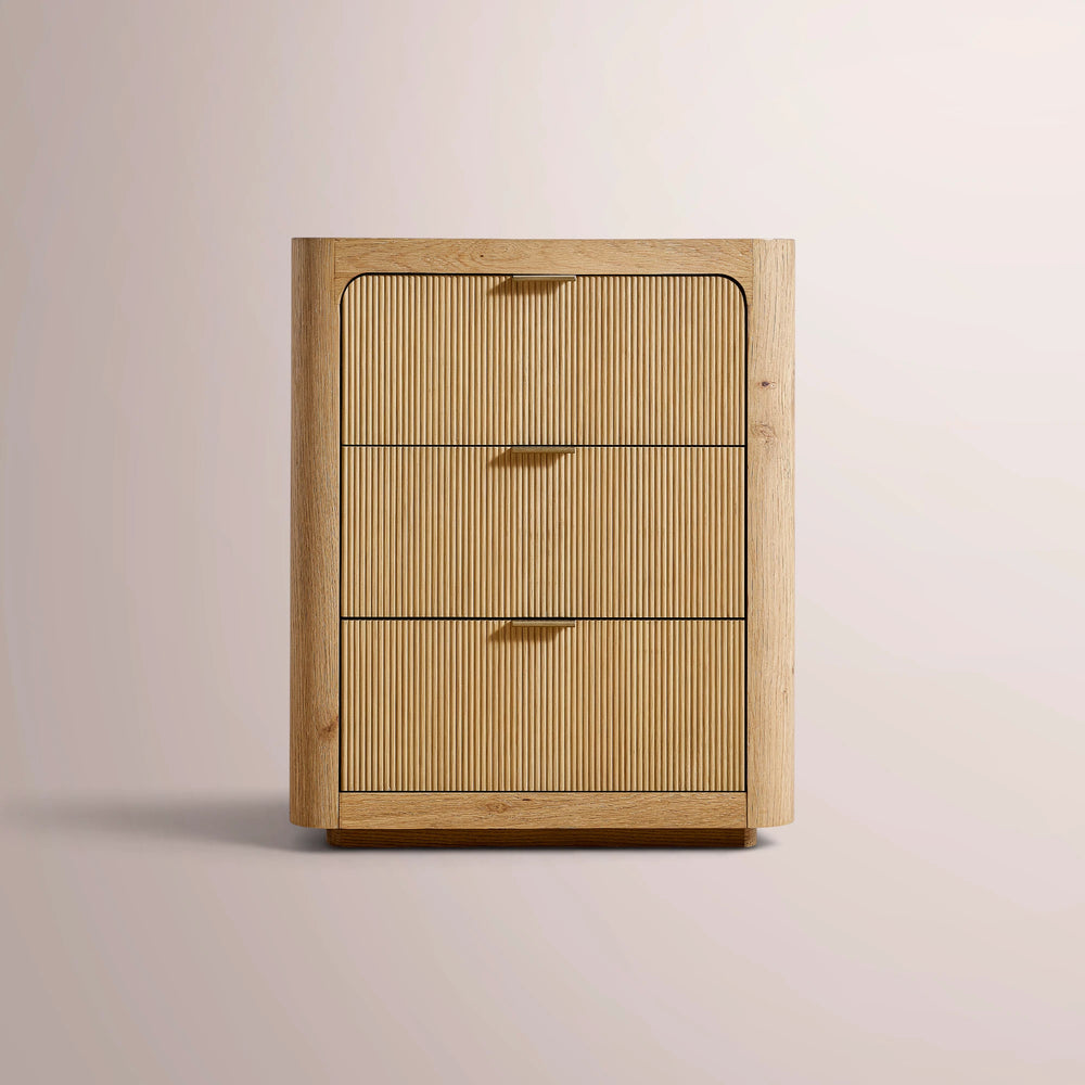 A three-drawer wooden chest or nightstand with a light oak finish, featuring vertically ribbed drawer fronts and small brass handles, set against a warm beige background.