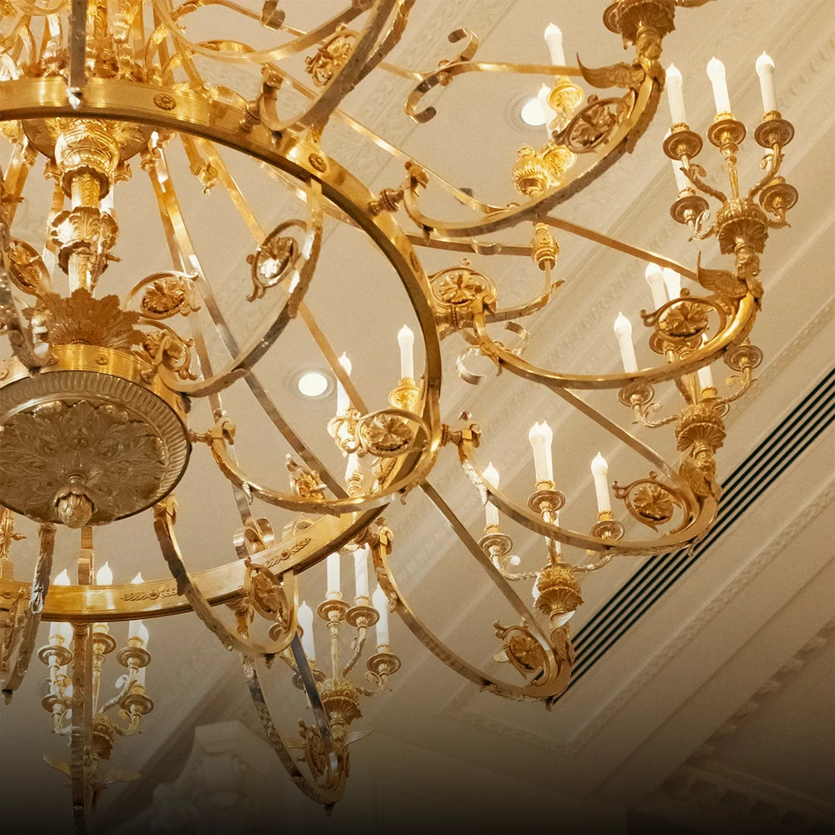 An ornate gold chandelier with multiple tiers, curved arms, and lit candle-style bulbs hanging from a decorated ceiling.