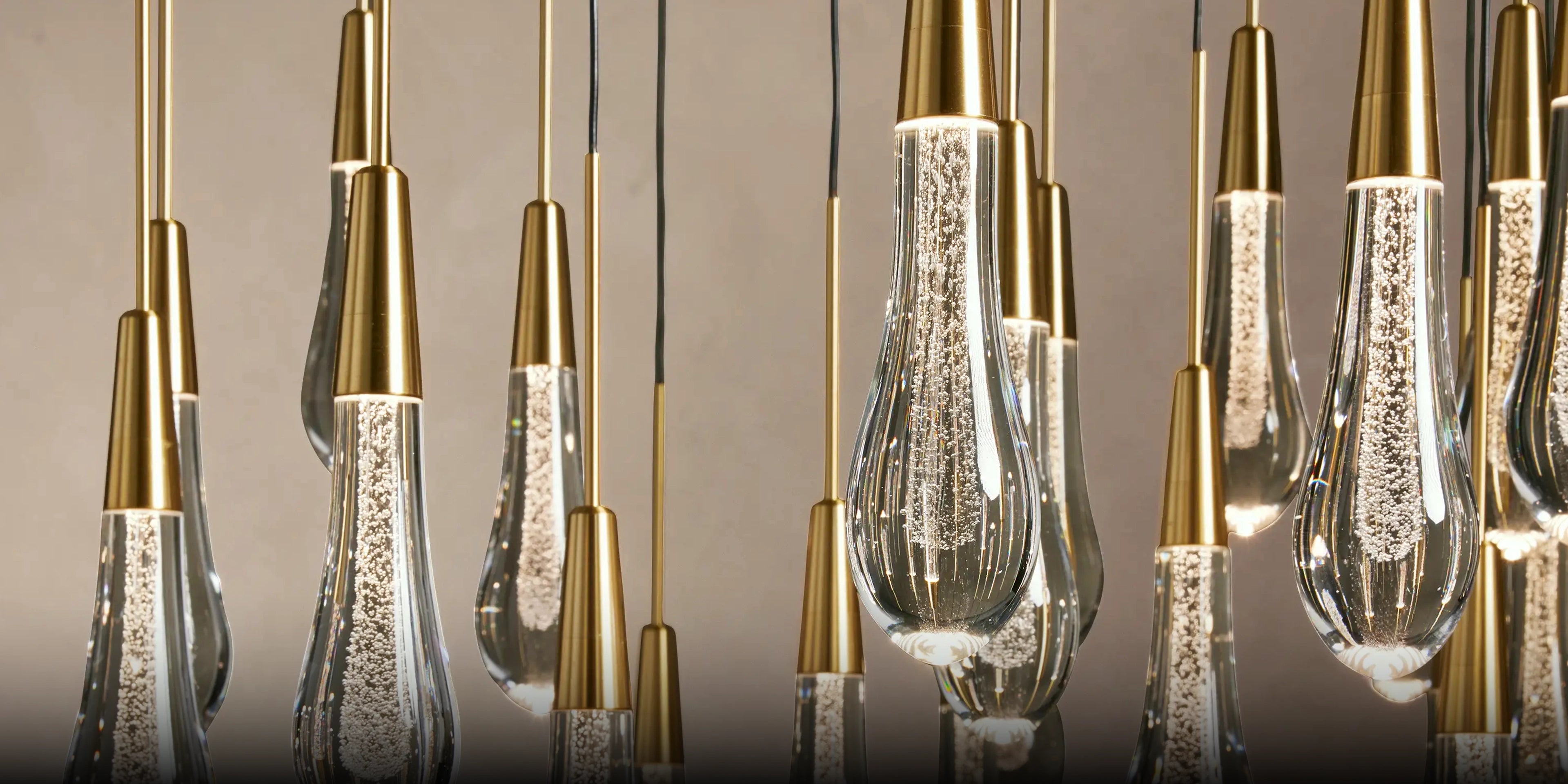 A close-up of a modern chandelier with multiple hanging glass teardrop-shaped bulbs with gold metal caps and clear textured glass bodies, shown against a warm neutral background.