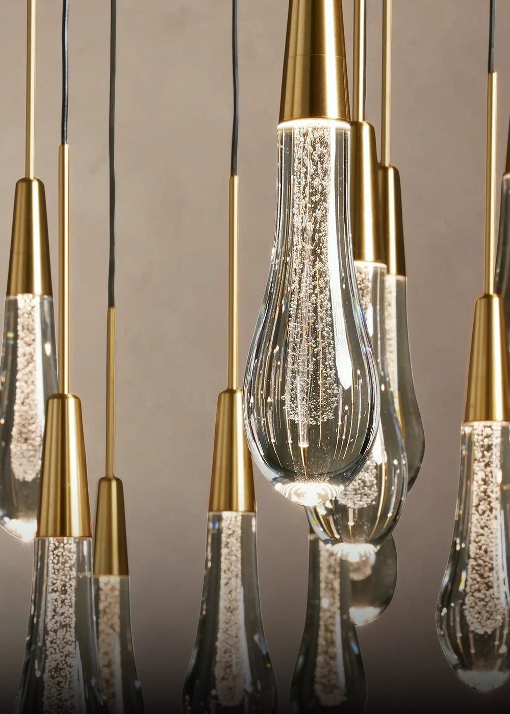 A close-up of a modern multi-light pendant chandelier with clear glass teardrop-shaped bulbs filled with suspended gold glitter, suspended from slim gold rods against a neutral gray background.